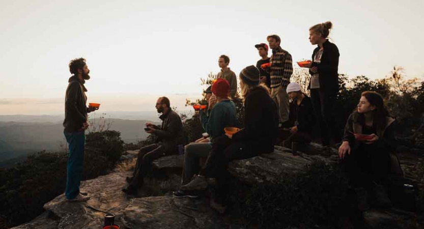 A group of people sit on a rocky overlook, listening to one of them speak. They appear to have bowls of food. 